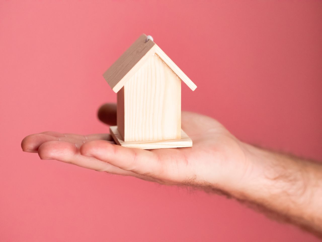 Mastering the First Impression: Your intriguing post title goes here Wooden house model in hand symbolizing real estate concepts on pink background.